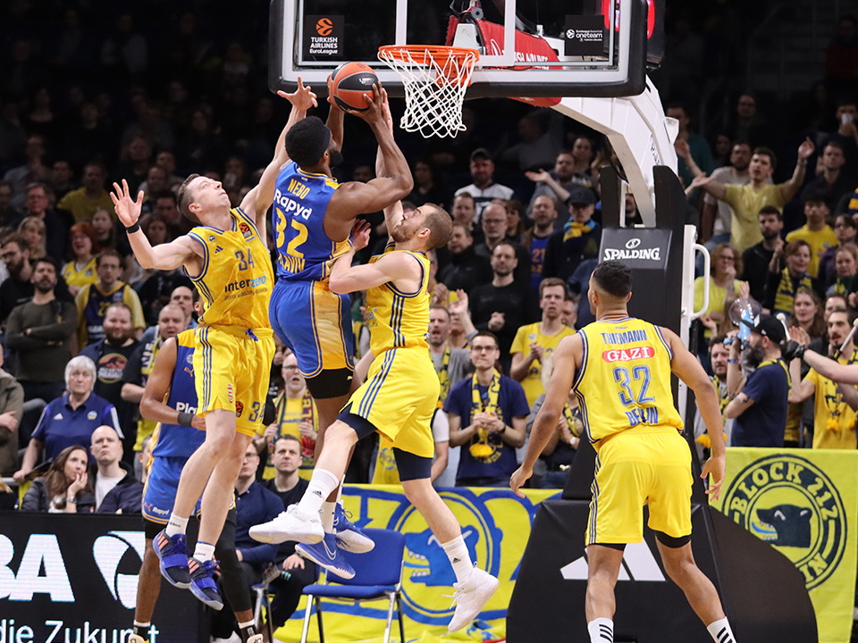 Image of a basketball game. A blue player attempts to shoot the ball into the basket, but two yellow players block him in the air. One player from each team watches closely. In the background, there is a full grandstand with people watching.